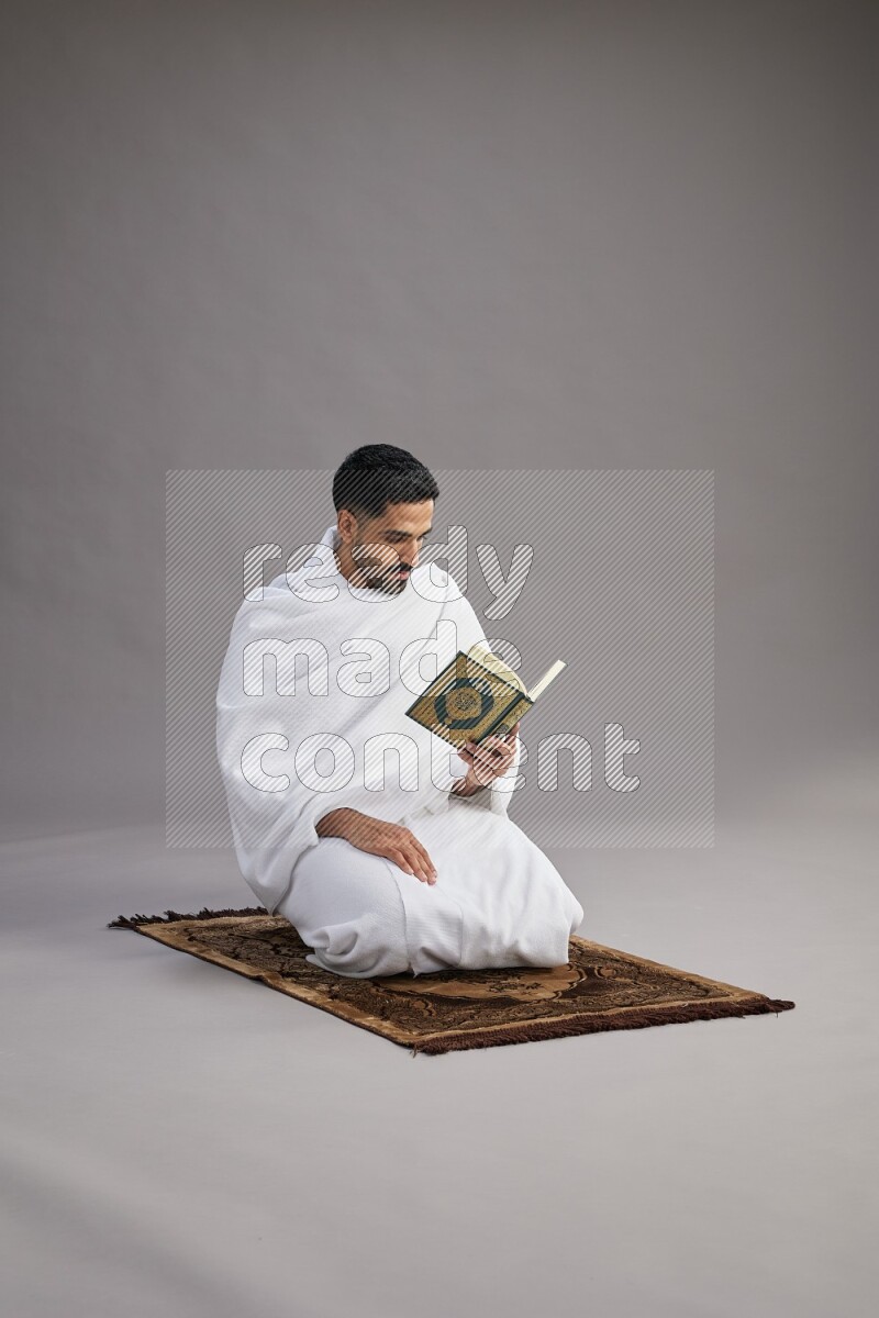 A man wearing Ehram sitting on floor reading quran on gray background