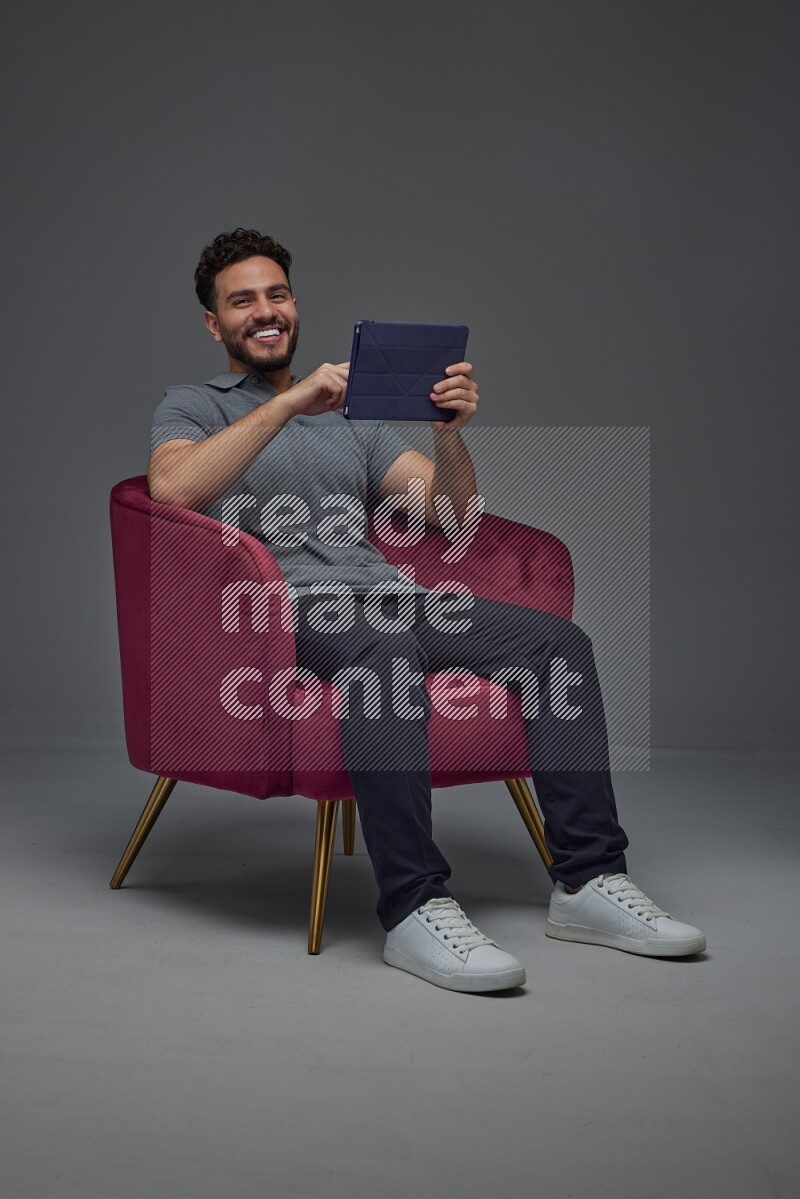 A man wearing casual and using his tablet while sitting on a burgundy chair eye level on a gray background