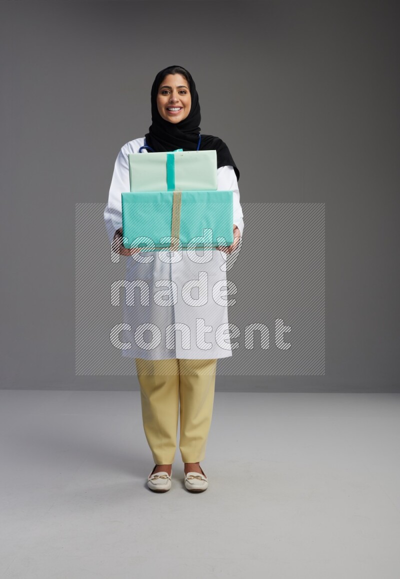 Saudi woman wearing lab coat with stethoscope standing holding gift box on Gray background