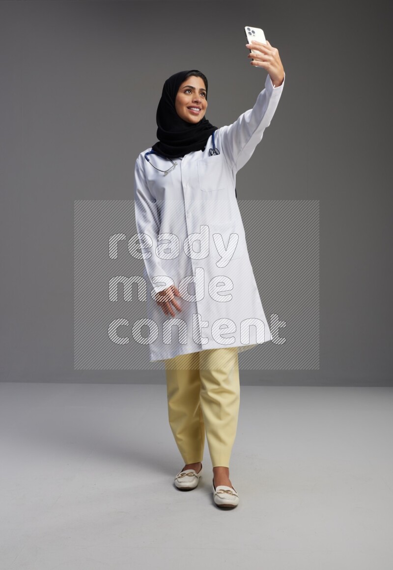 Saudi woman wearing lab coat with stethoscope standing taking selfie on Gray background