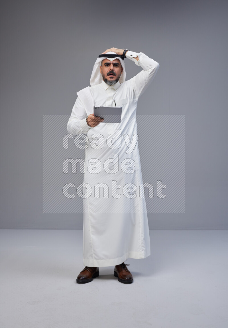 Saudi man Wearing Thob and white Shomag standing working on tablet on Gray background
