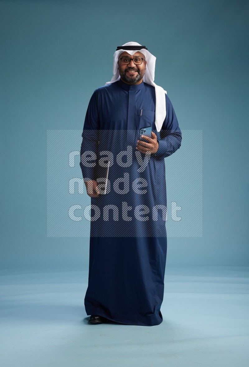 Saudi Man with shimag Standing texting on phone on blue background