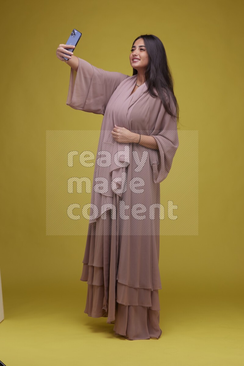 A woman Taking a Selfie on a Yellow Background wearing Brown Abaya
