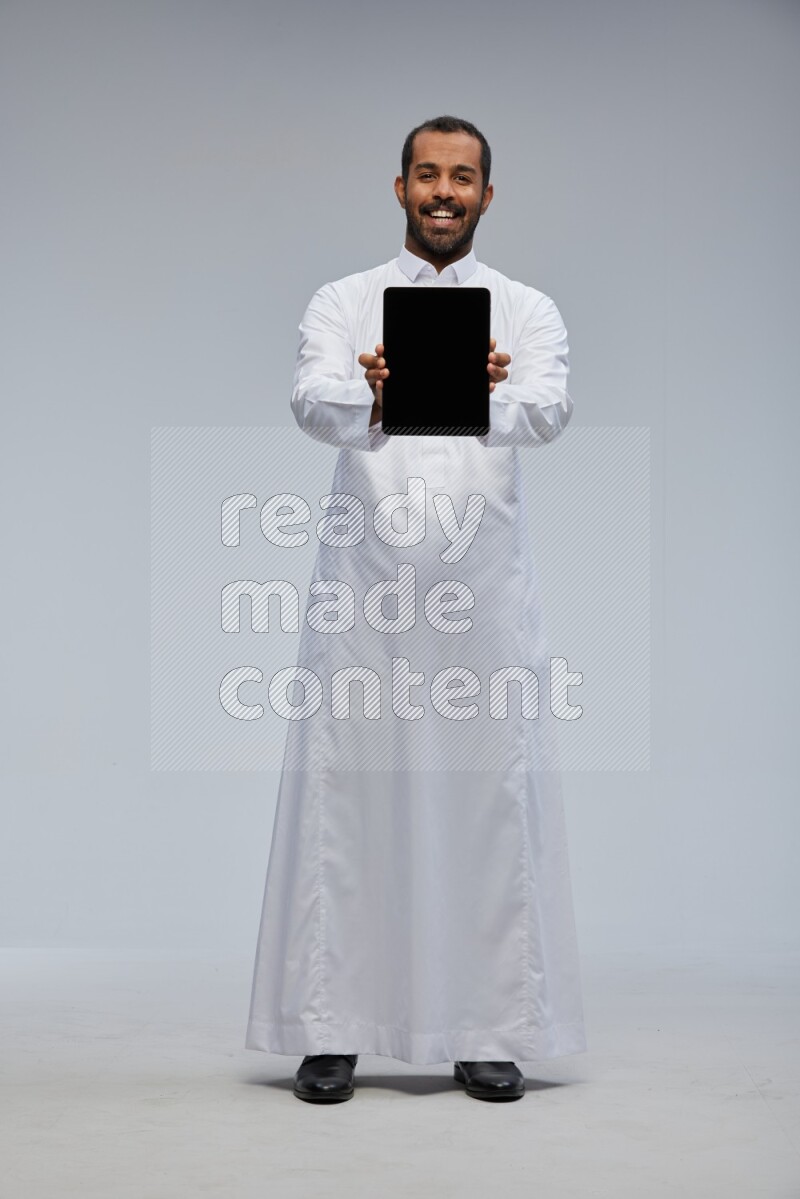 Saudi man wearing Thob standing showing tablet to camera on Gray background