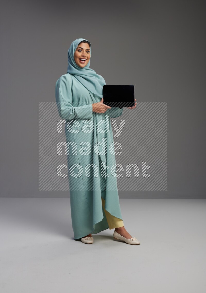 Saudi Woman wearing Abaya standing showing tablet to camera on Gray background
