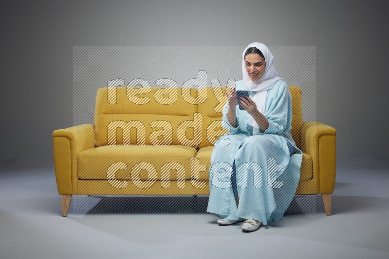 A Saudi woman wearing a light blue Abaya and white head scarf sitting on a yellow sofa and using her phone eye level on a grey background