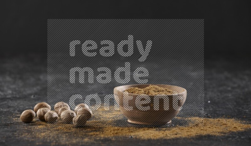 A wooden bowl full of nutmeg powder with whole seeds and sprinkled powder beside it on a textured black flooring