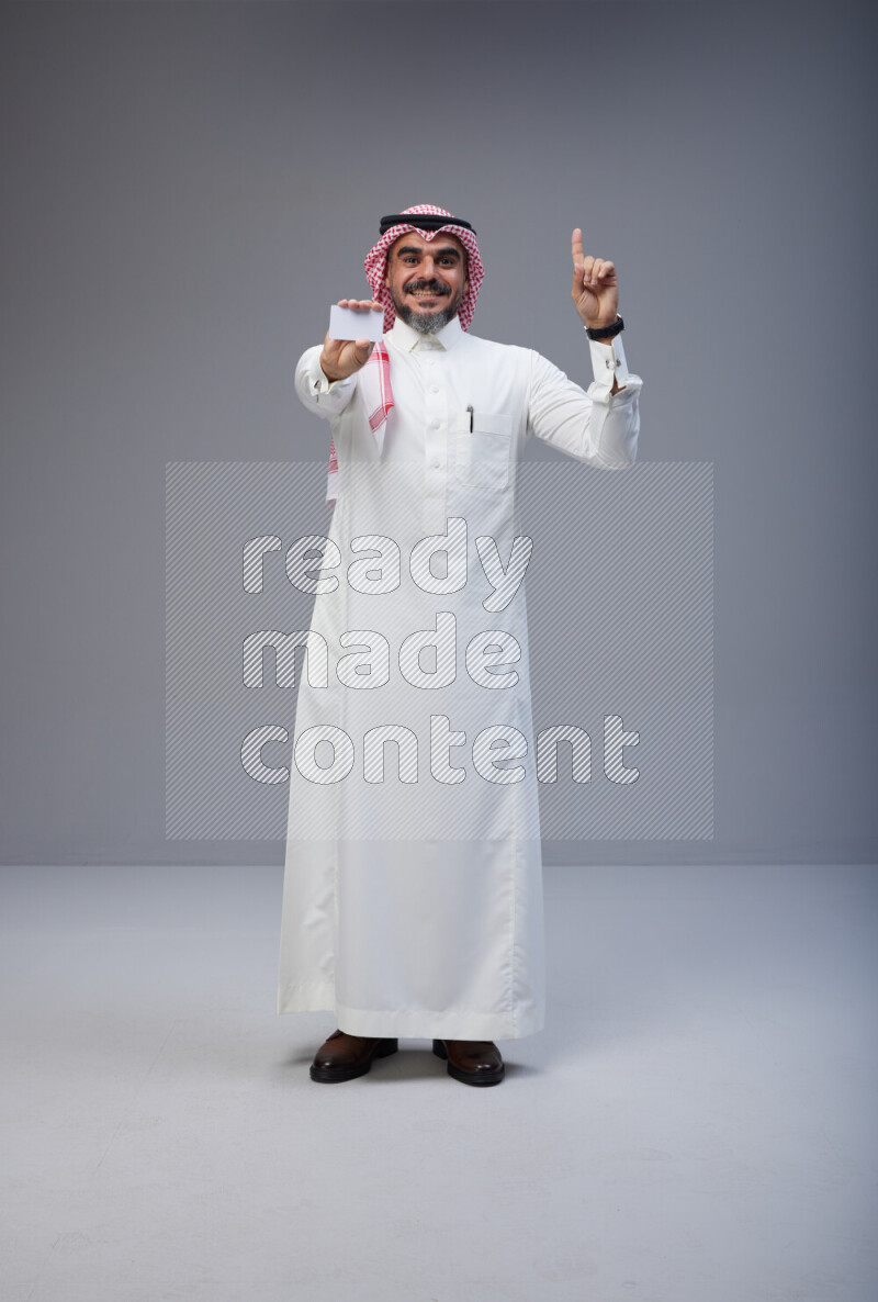 Saudi man Wearing Thob and red Shomag standing holding ATM card on Gray background