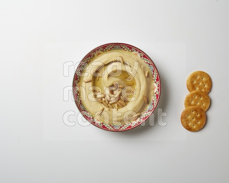 Hummus in a red plate with patterns garnished with pine nuts on a white background
