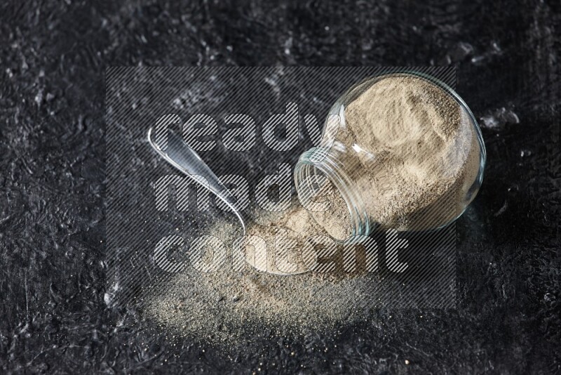A flipped herbal glass jar and a metal spoon full of white pepper powder with spilled powder on textured black flooring