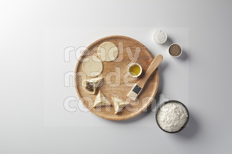 two closed sambosas and one open sambosa filled with meat while flour, salt, black pepper and oil with oil brush aside in a wooden dish on a white background