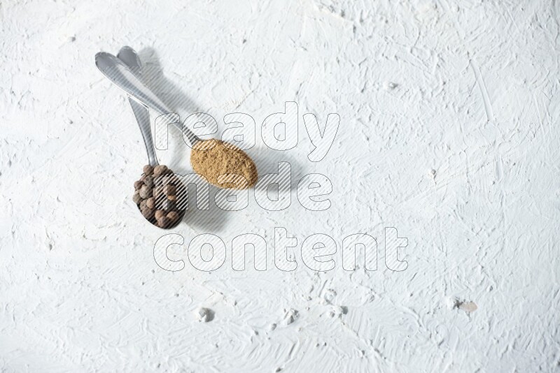 2 metal spoons full of allspice powder and whole balls on a textured white flooring