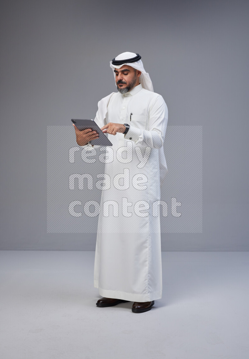 Saudi man Wearing Thob and white Shomag standing working on tablet on Gray background