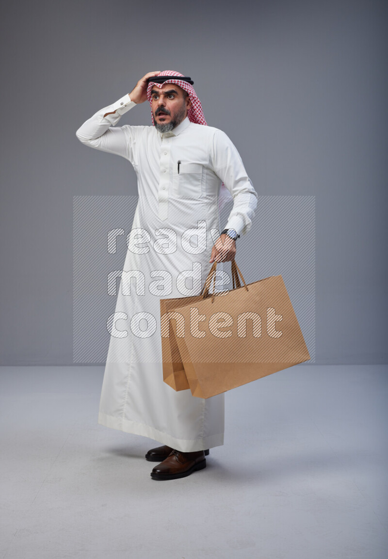 Saudi man Wearing Thob and red Shomag standing holding shopping bag on Gray background
