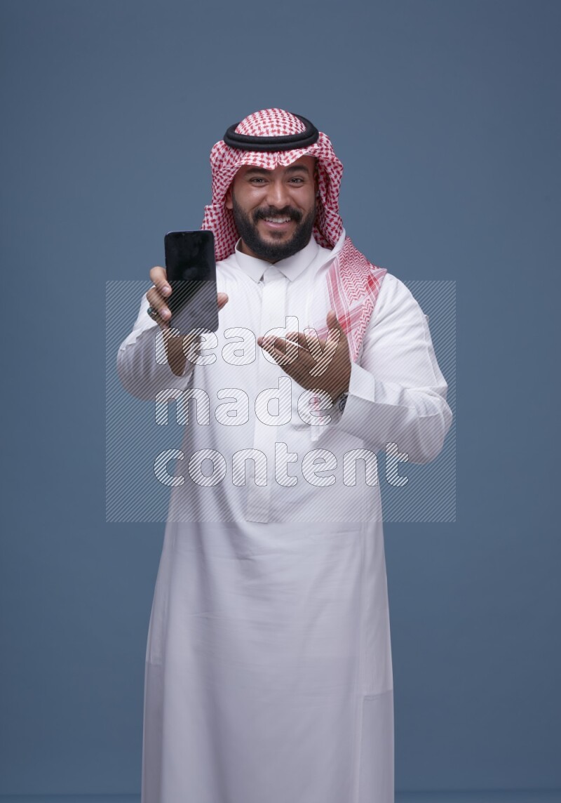 A man Showing his smartphone Screen on Blue Background wearing Saudi Thob