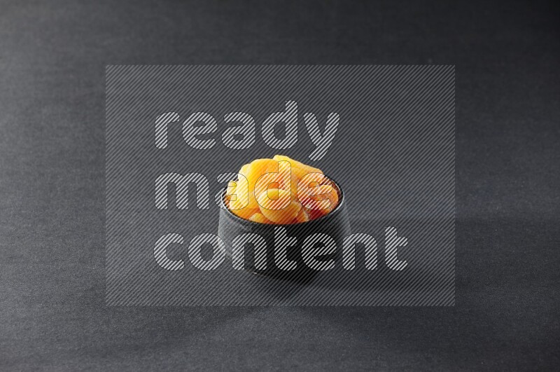 A black pottery bowl full of dried apricots on a black background in different angles