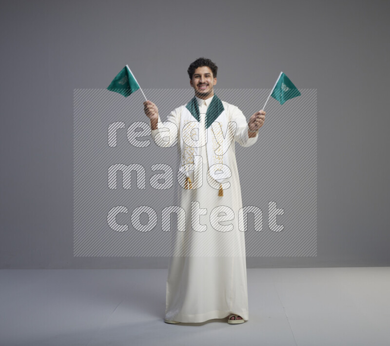 A Saudi man standing wearing thob and saudi flag scarf and holding small saudi flag on gray background