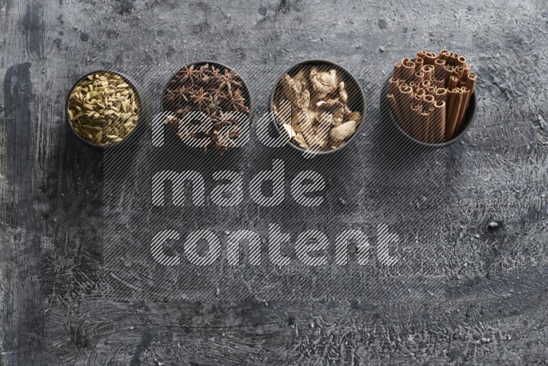 Ginger, Cardamom, Star anise and cinnamon sticks in 4 bowls on a textured black background