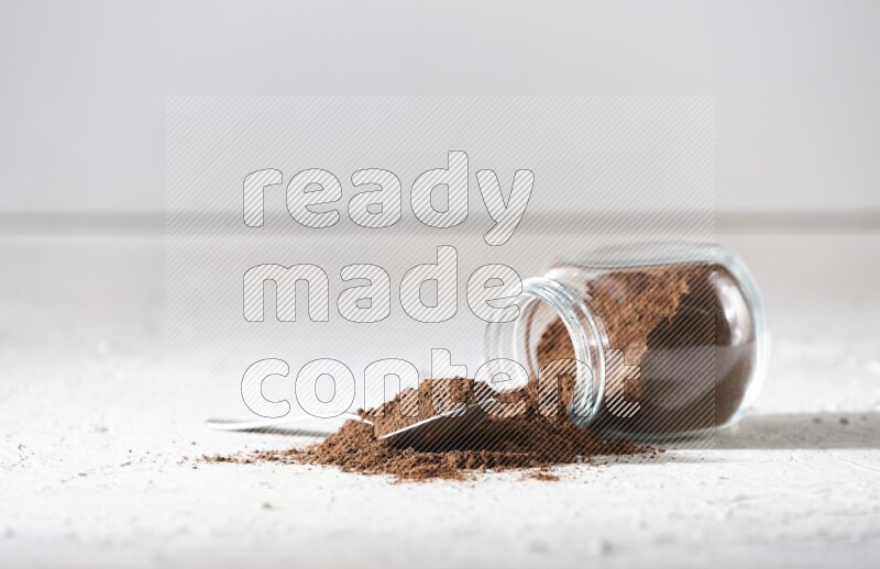 A flipped glass spice jar and a metal spoon full of cloves powder and powder came out of the jar on textured white flooring