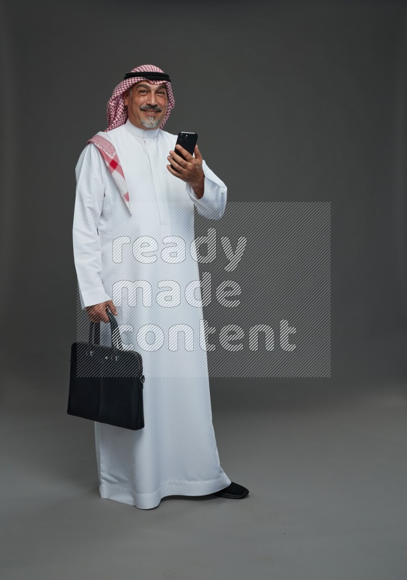 Saudi man with shomag Standing holding bag and phone on gray background