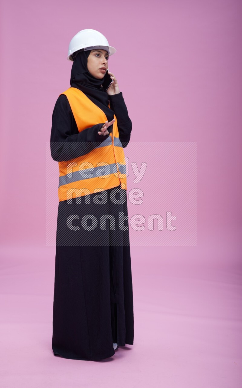 Saudi woman wearing Abaya with engineer vest and helmet standing talking on phone on pink background