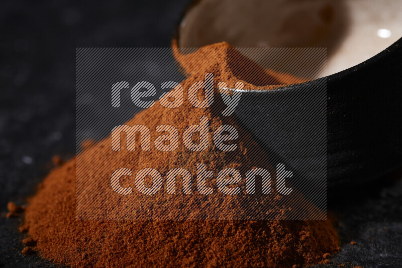 A black pottery bowl full of ground paprika powder with fallen powder from it on black background