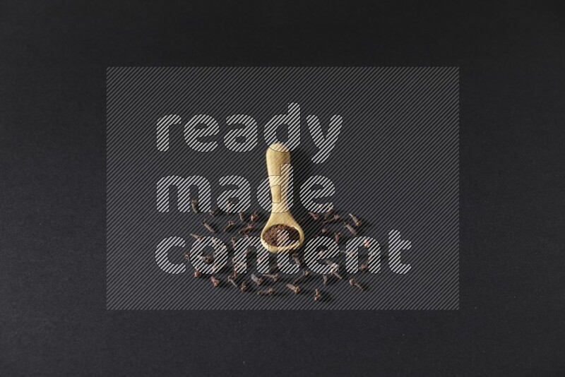 ملعقة خشبية ممتلئة ببودرة القرنفل مع حبوب القرنفل المتناثرة على خلفية سوداء