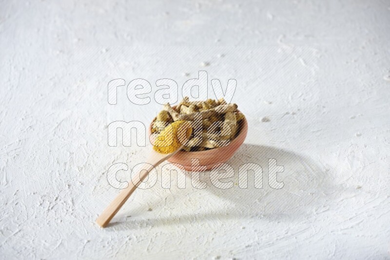 A wooden spoon full of turmeric powder above a wooden bowl full of dried turmeric whole fingers on a textured white flooring
