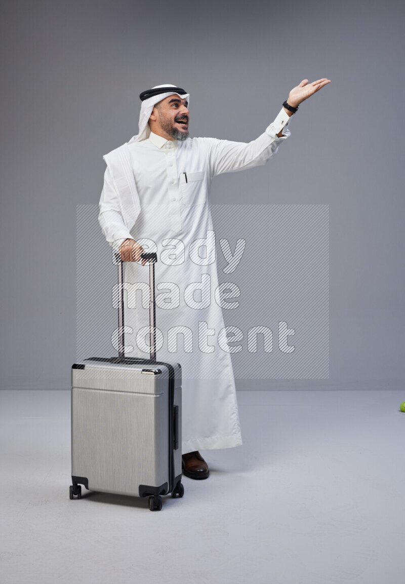 Saudi man wearing Thob and white Shomag standing holding Travel bag on Gray background