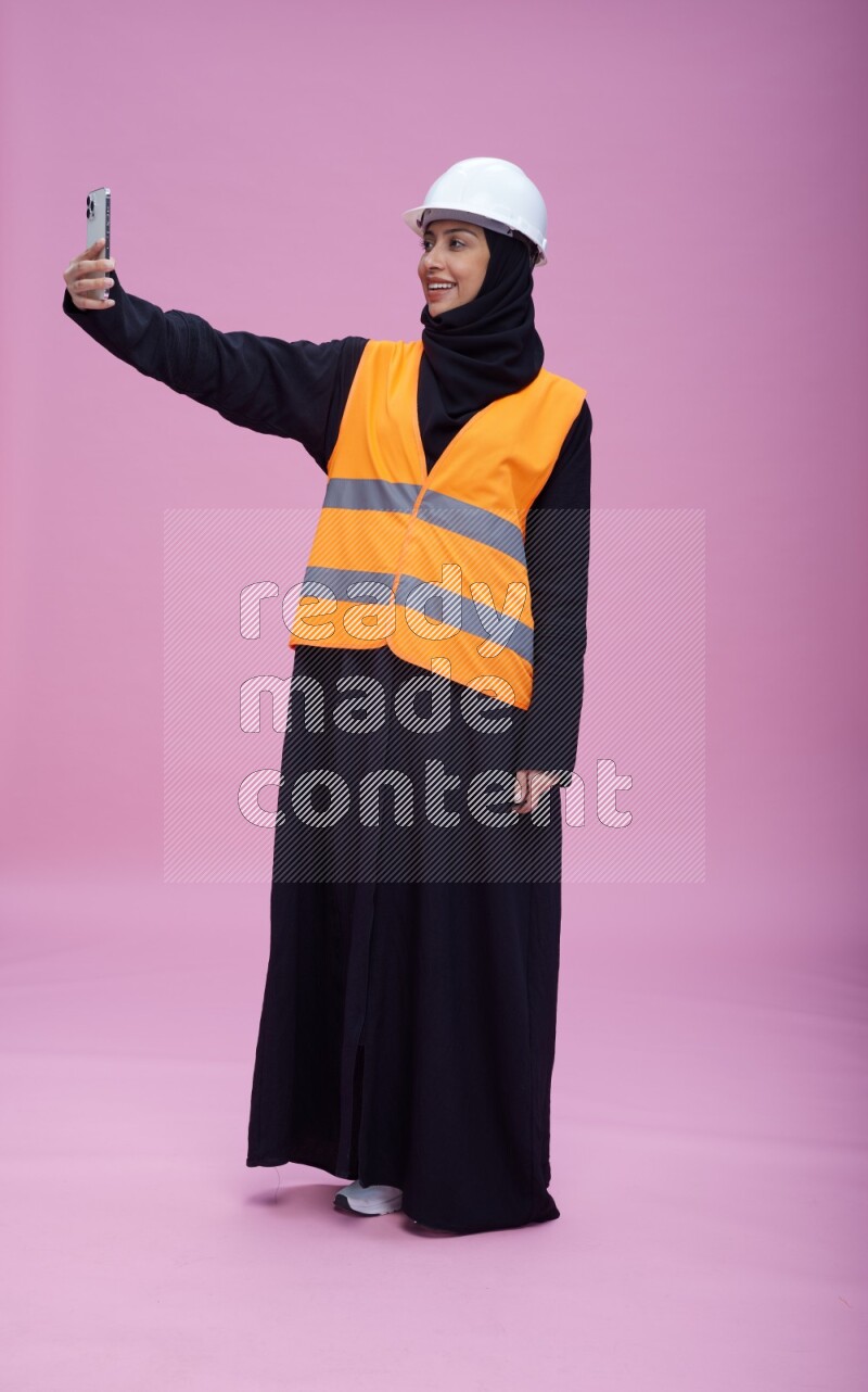 Saudi woman wearing Abaya with engineer vest and helmet standing taking selfie on pink background