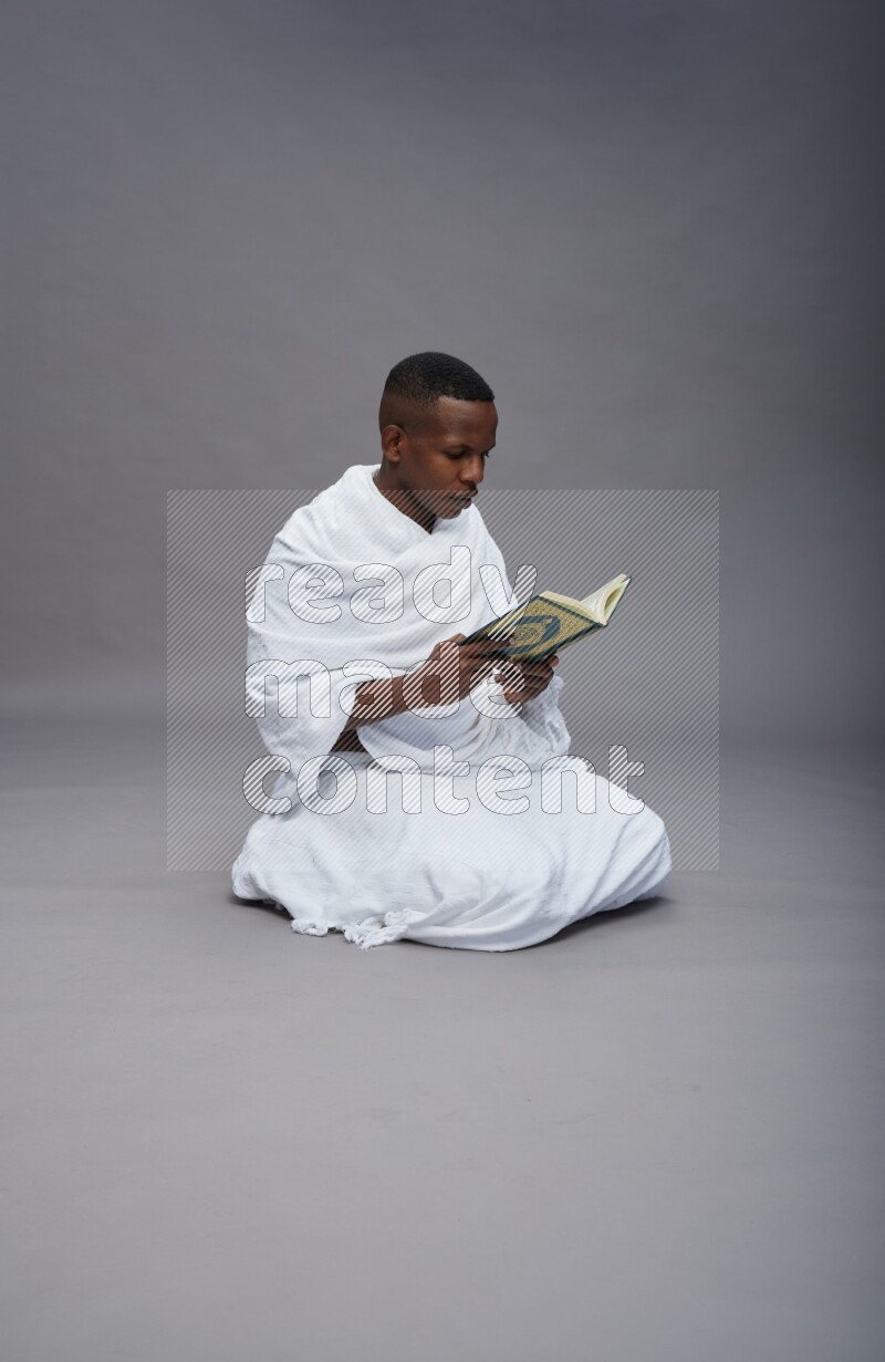 A man wearing Ehram sitting on floor reading quran on gray background