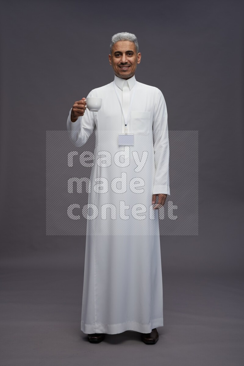 Saudi man wearing thob with neck strap employee badge standing holding mug on gray background