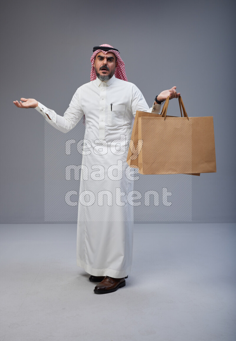 Saudi man Wearing Thob and red Shomag standing holding shopping bag on Gray background