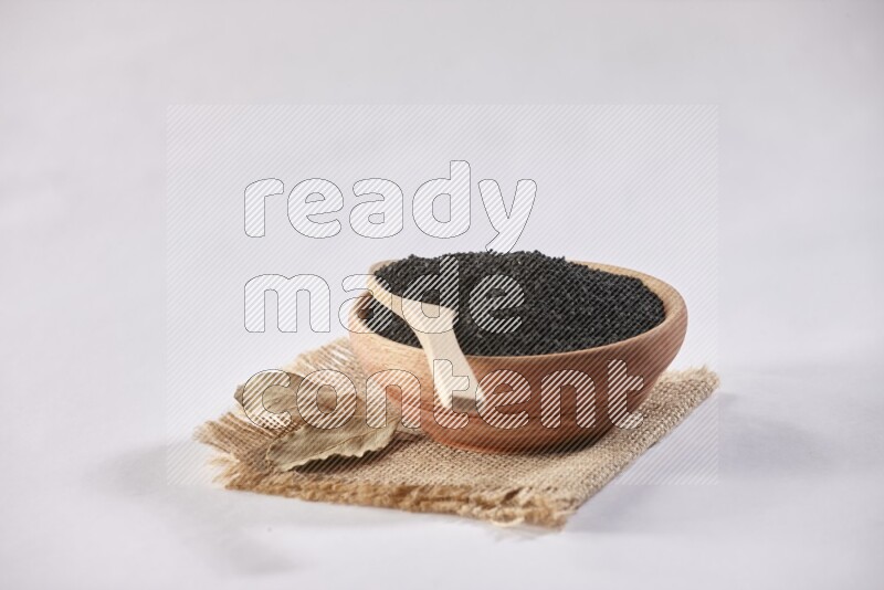 A wooden bowl and wooden spoon full of black seeds on a piece of burlap on a white flooring