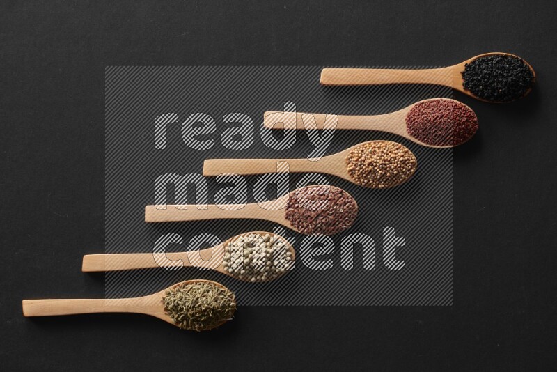 wooden spoons filled with white peppers, mustard seeds, black seeds, garden cress, cumin and flax on black flooring