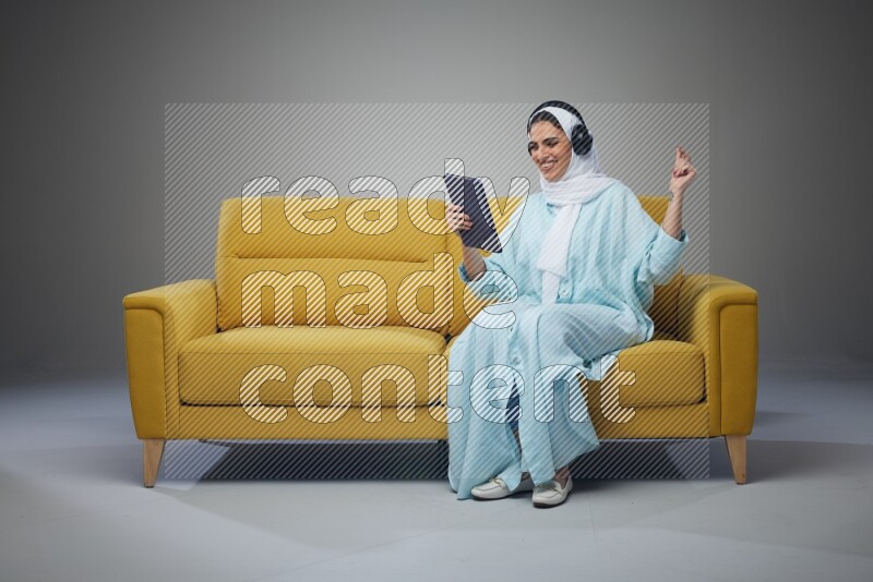 A Saudi woman wearing a light blue Abaya and a white head scarf sitting on a yellow sofa and holding an iPad while wearing headphone eye level on a grey background