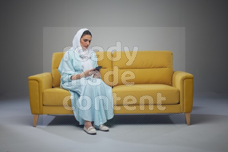 A Saudi woman wearing a light blue Abaya and white head scarf sitting on a yellow sofa and using her phone eye level on a grey background