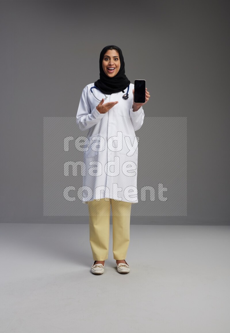 Saudi woman wearing lab coat with stethoscope standing showing phone to camera with sign in the back on Gray background