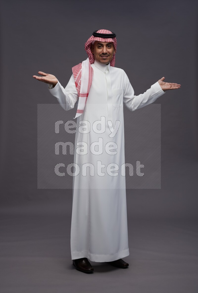 Saudi man wearing thob and shomag standing interacting with the camera on gray background