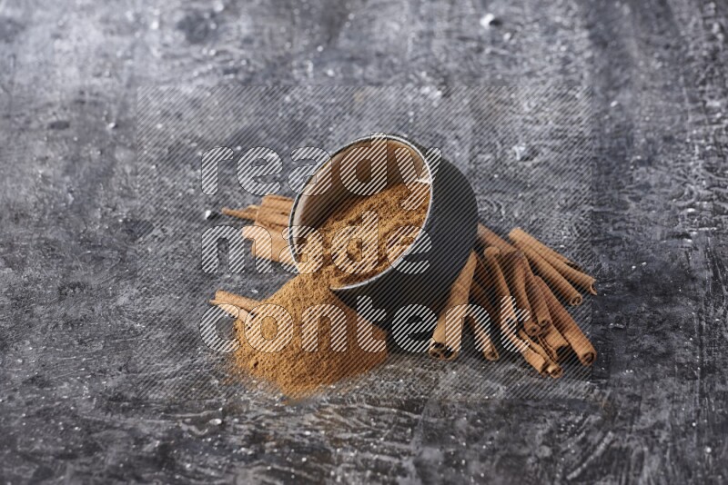 Black pottery bowl over filled with cinnamon powder and cinnamon sticks around the bowl on a textured black background