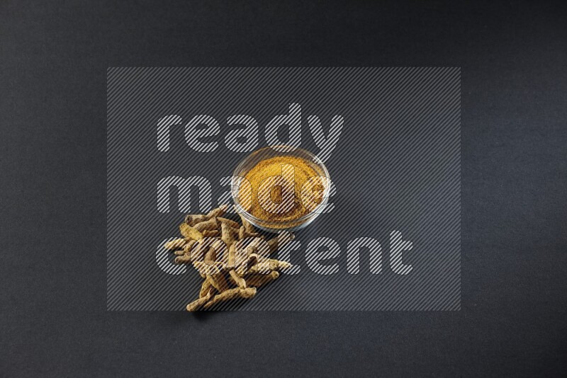 A glass bowl full of turmeric powder and dried whole fingers beside it on black flooring