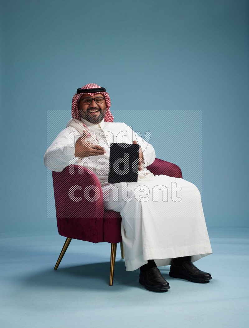 Saudi Man with shimag sitting on chair working on tablet on blue background