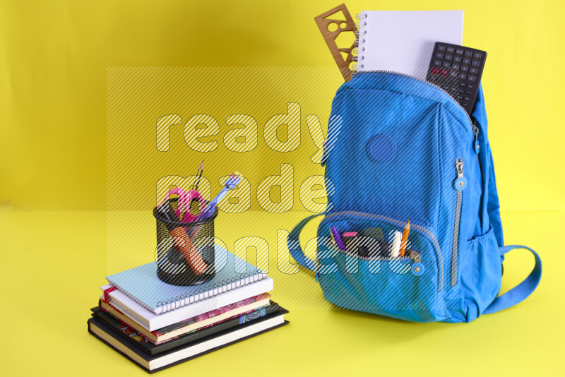 A school bag with assorted school supplies in and beside it on yellow background