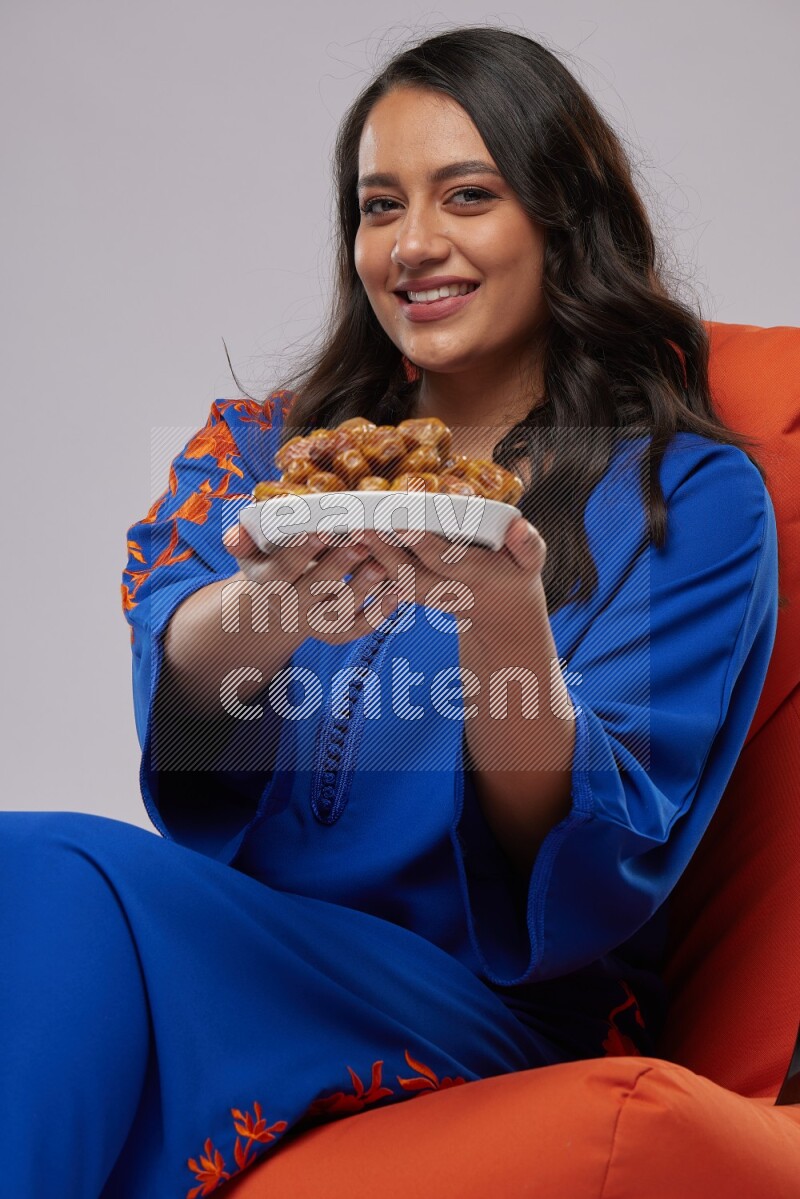 A Woman sitting on an orange beanbag wearing Jalabeya holding a plate of dates
