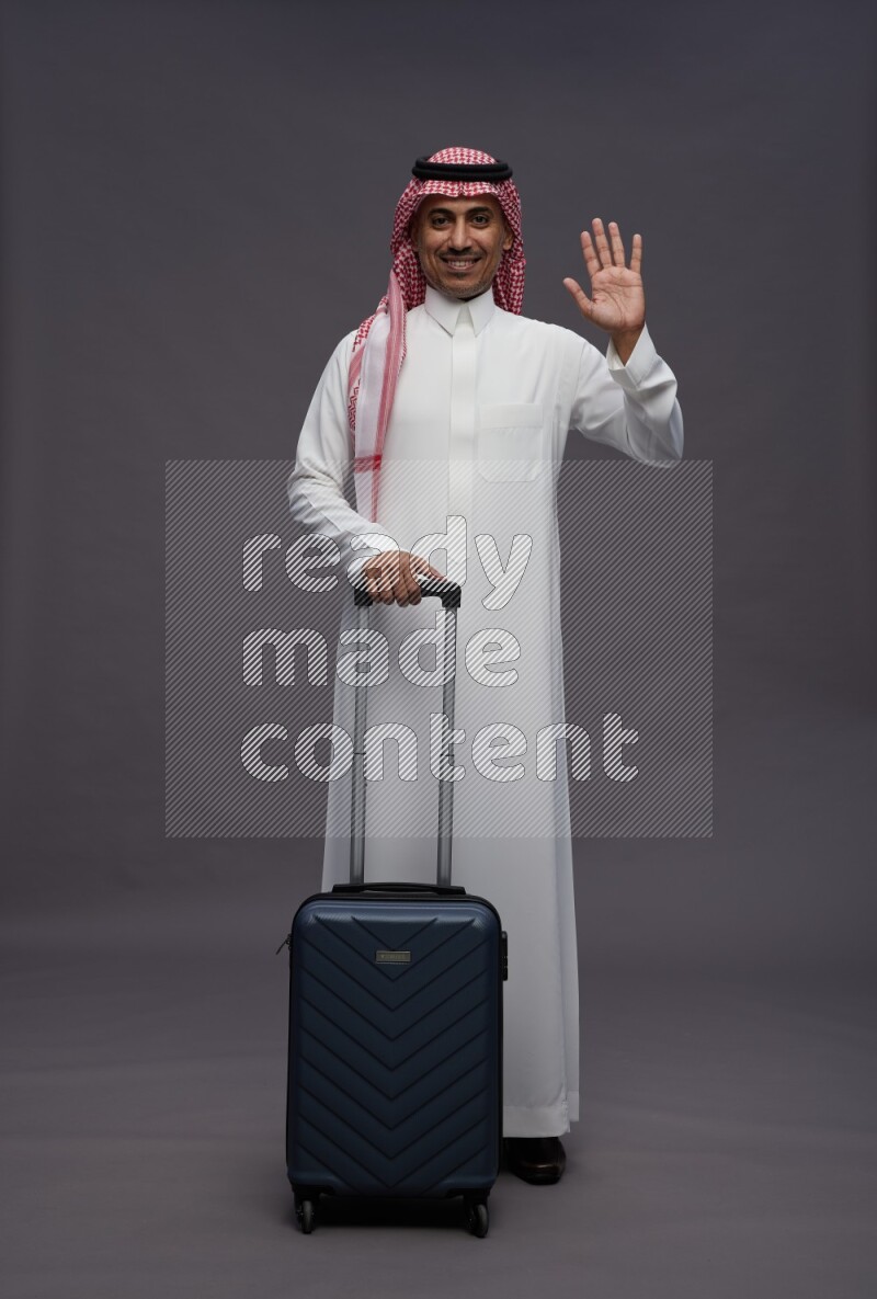 Saudi man wearing thob and shomag standing holding bag on gray background