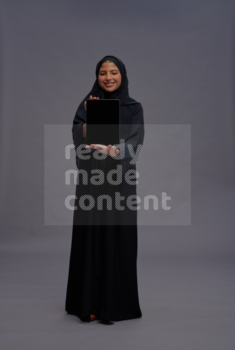 Saudi woman wearing Abaya standing showing tablet to camera on gray background