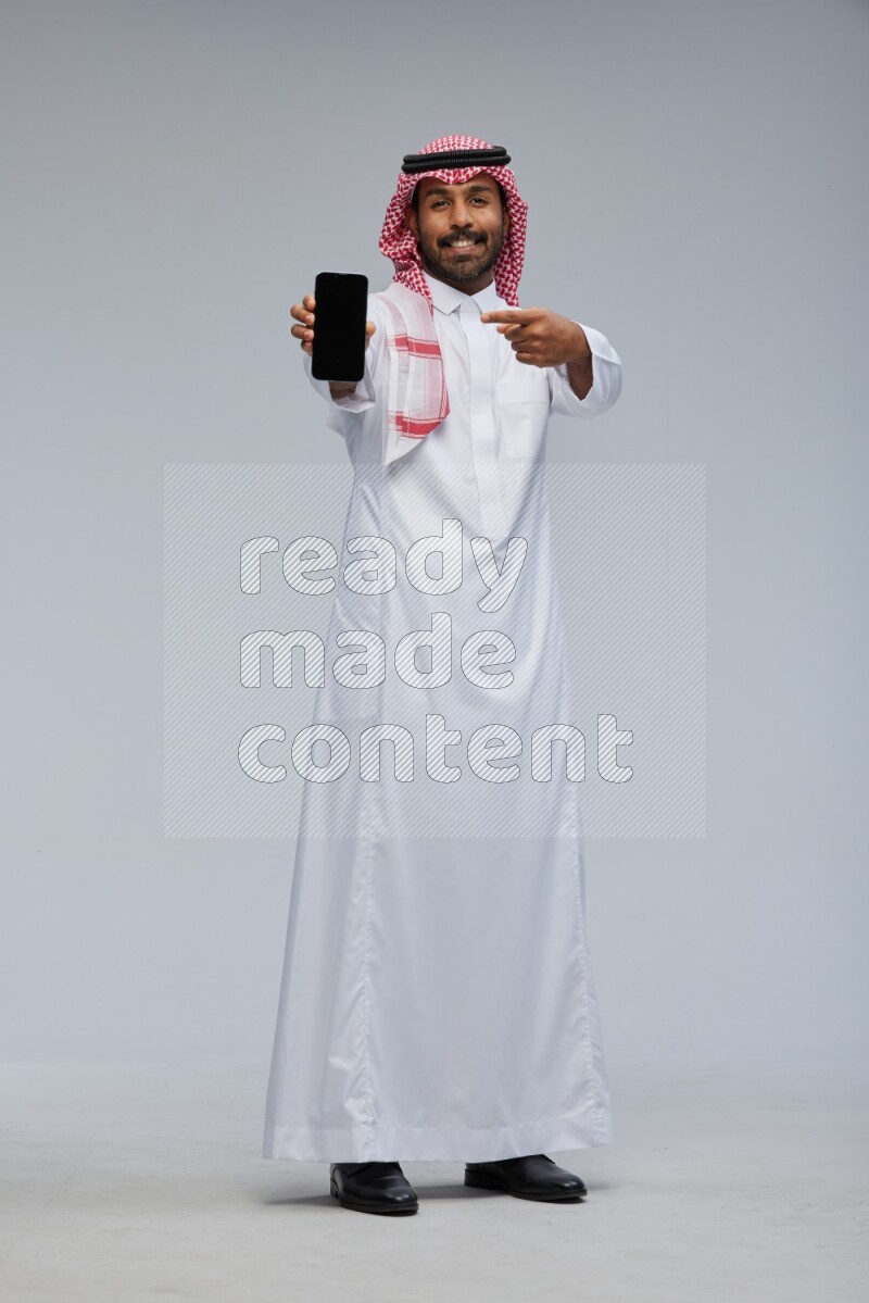 Saudi man Wearing Thob and shomag standing showing phone to camera on Gray background