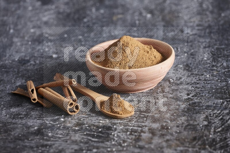 wooden bowl full of cinnamon powder and a wooden spoon full of it with cinnamon sticks on a textured black background