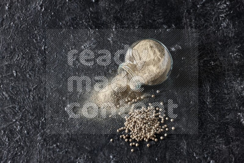 A flipped herbal glass jar full of white pepper powder with spilled powder and pepper beads on textured black flooring
