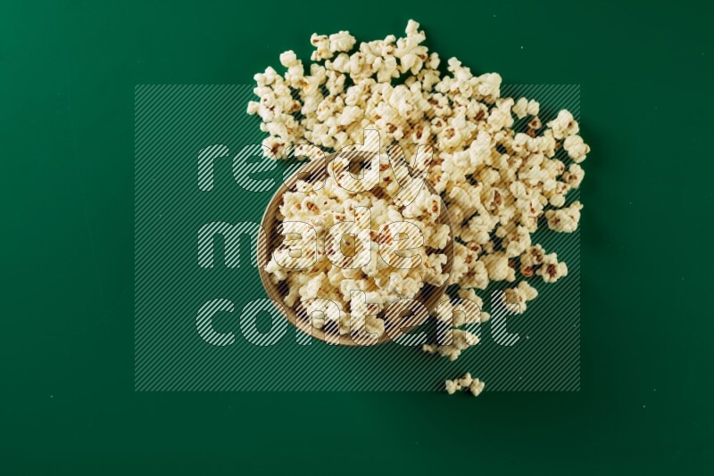 An off white ceramic bowl full of popcorn with popcorn beside it on a green background in a top view shot